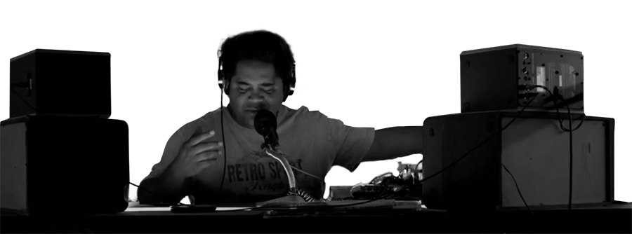 Imagem em preto e branco de homem negro com corte de cabelo arredondado sentado atrás de equipamentos de som e um microfone.