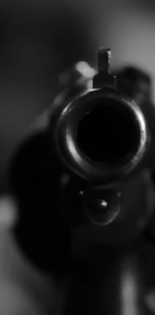 Imagem em preto e branco de um cano de um revólver, em primeiro plano, apontado em direção à câmera.
