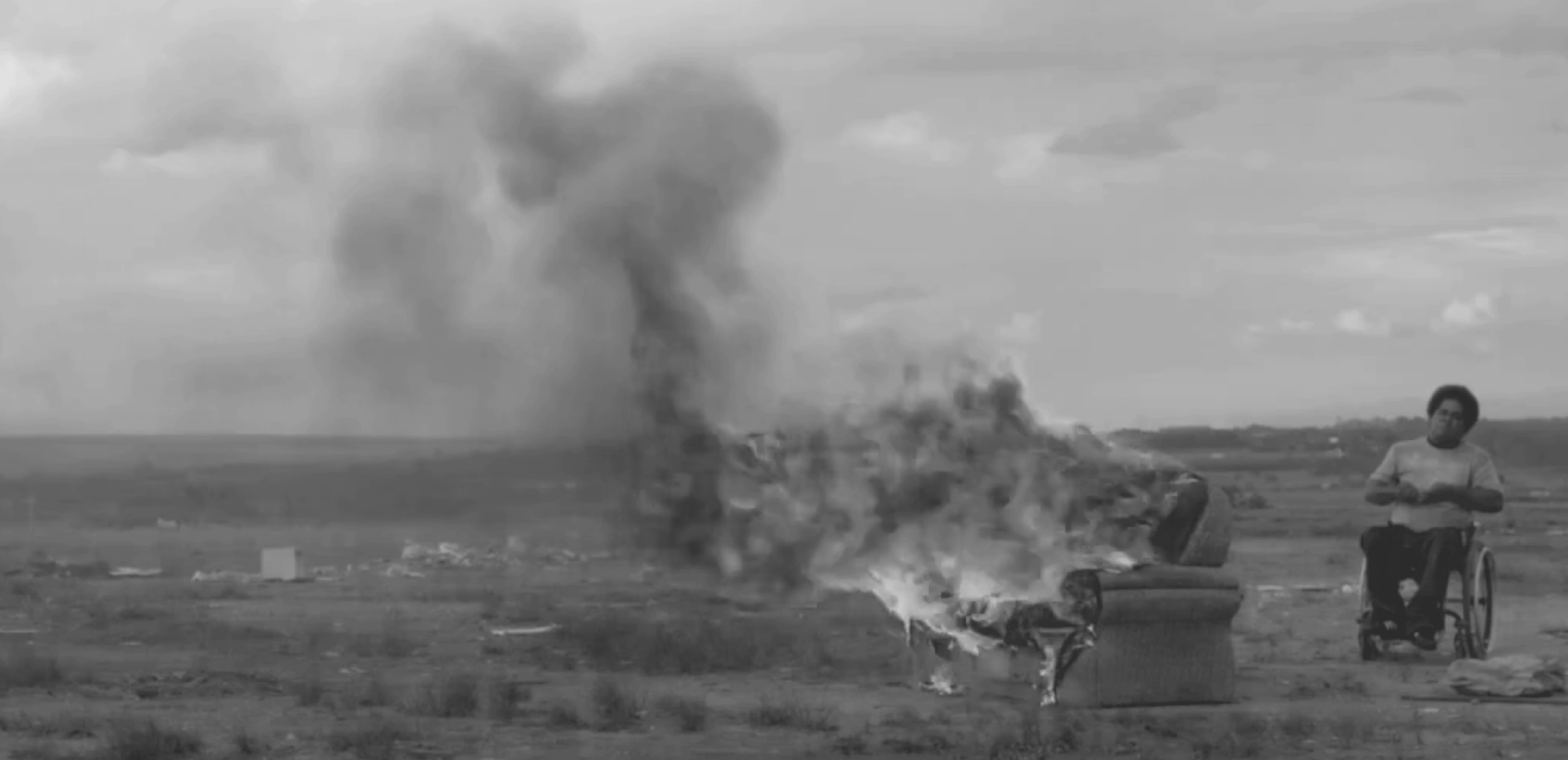 Imagem em preto e branco de homem negro cadeirante, de cabelo com corte arrendodado, olhando para um sofá pegando fogo.